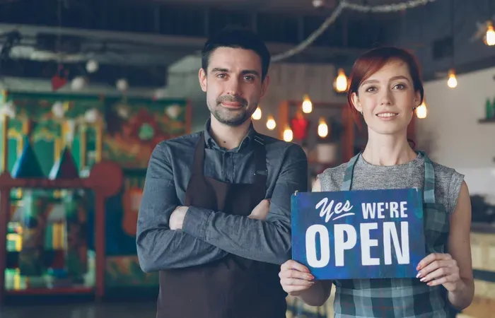 Two restaurant owners smiling and holding a “Yes, We’re Open” sign inside their warmly lit restaurant on localservicemap.net